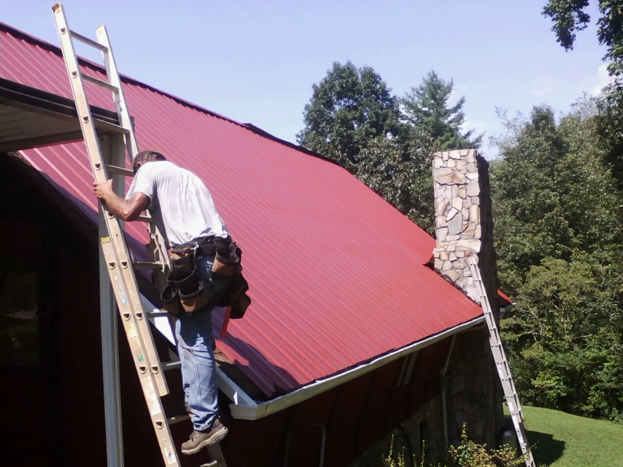 Roofer on a ladder beside a red metal roof and stone chimney — Soffit & Fascia Repair by Roofing Brothers in Byars